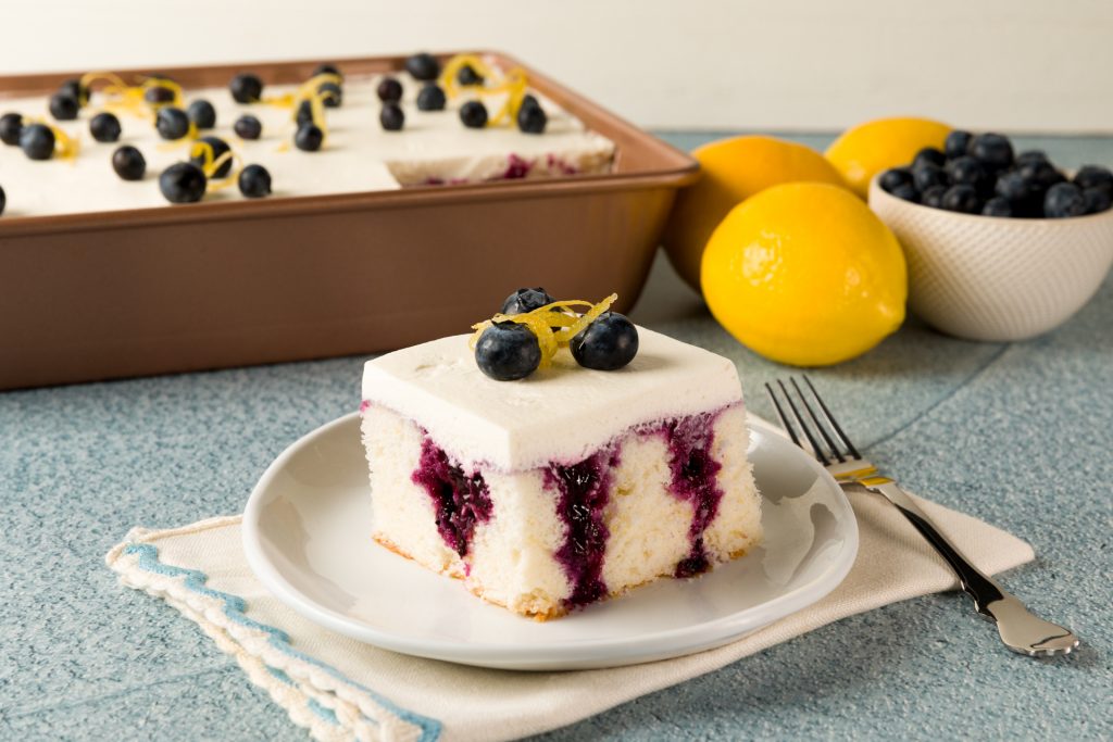 Slice of Lemon Berry Poke Cake with a lemon and the rest of the cake in the background